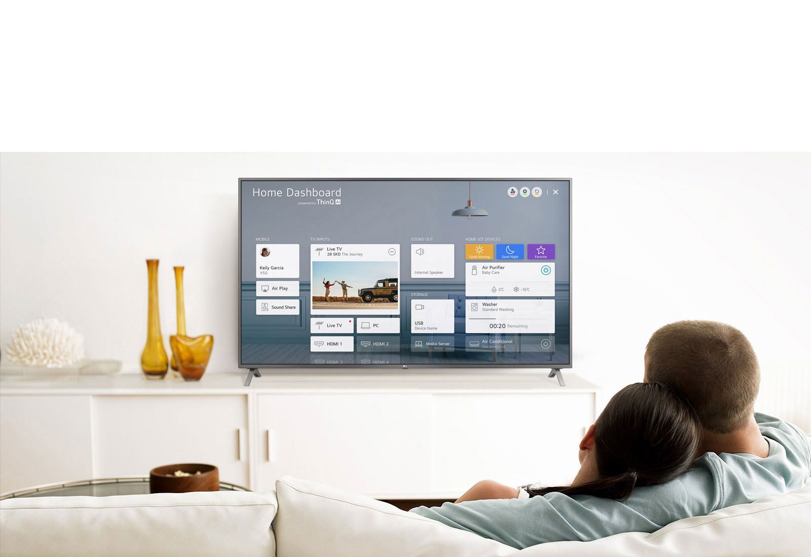 A men and women sitting on a sofa in the living room with the Home Dashboard on the TV screen.