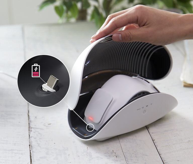 A hand holds open the lid of the purifier case to show the LG PuriCare Wearable Air Purifier sitting inside. There is a red light on the side of the wearable air purifier inside and inset image in a circle with a line to the side of the mask shows a magnified view of the place to recharge the mask. There is also a battery icon that is red and almost empty indicating it's time to plug it in.