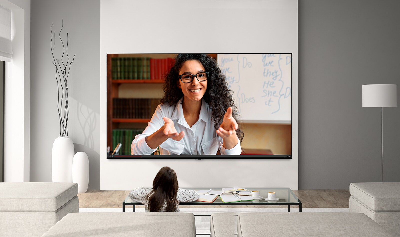 A young girl sat studying at a coffee table while watching a video lecture on a large, wall-mounted LG QNED Mini LED TV.