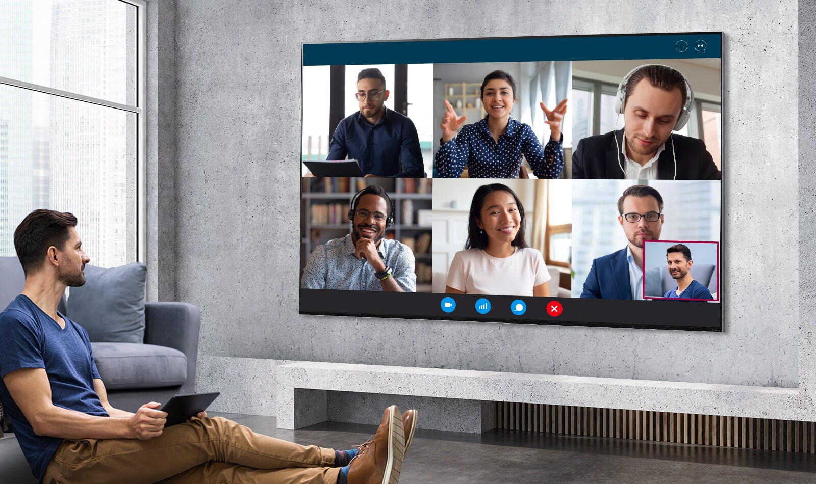 A man sat on the floor holding a tablet pc and watching a video meeting on a large, wall-mounted LG QNED Mini LED TV.