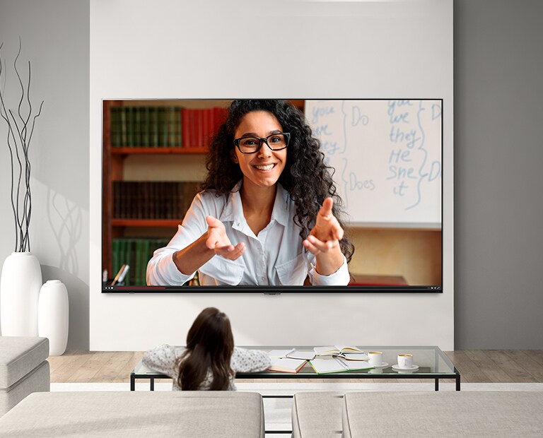 A young girl sat studying at a coffee table while watching a video lecture on a large, wall-mounted LG QNED Mini LED TV.