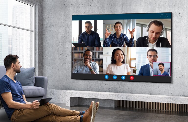 A man sat on the floor holding a tablet pc and watching a video meeting on a large, wall-mounted LG QNED Mini LED TV.