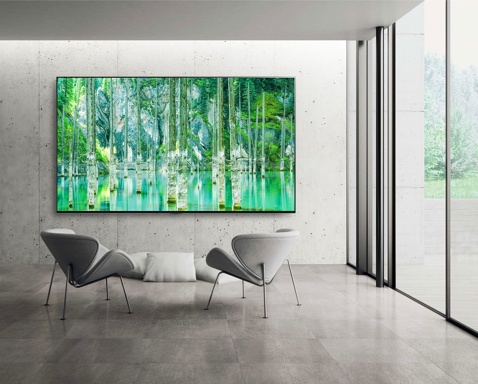 Image of a large-screen LG QNED MiniLED TV mounted against a grey, bare concrete wall. Two chairs are arranged in front of the TV and the screen shows a green natural scene.