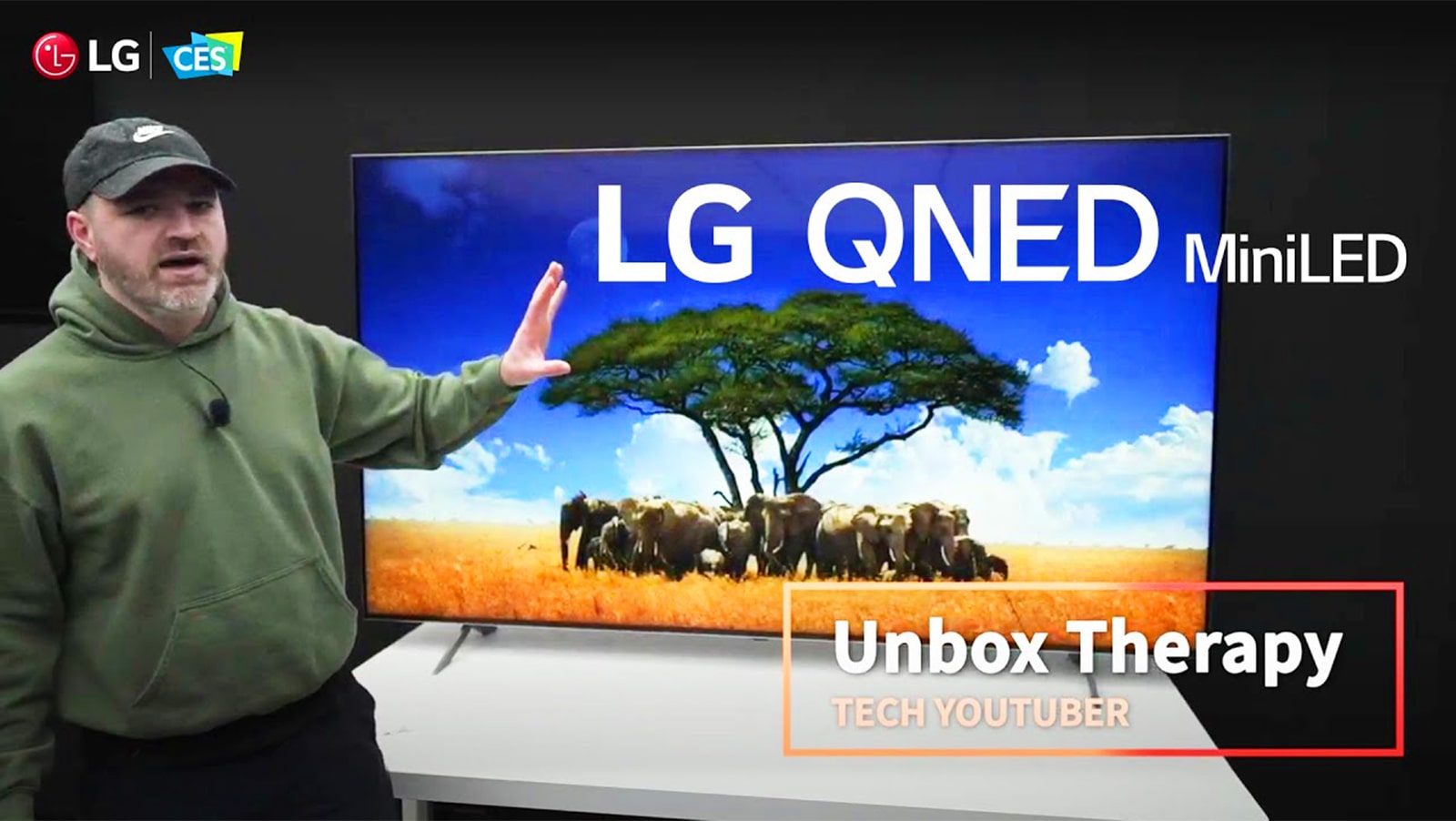 Tech YouTuber Unbox Therapy standing in front of an LG QNED MiniLED TV. The screen shows an image of a herd of elephants surrounding a single tree.