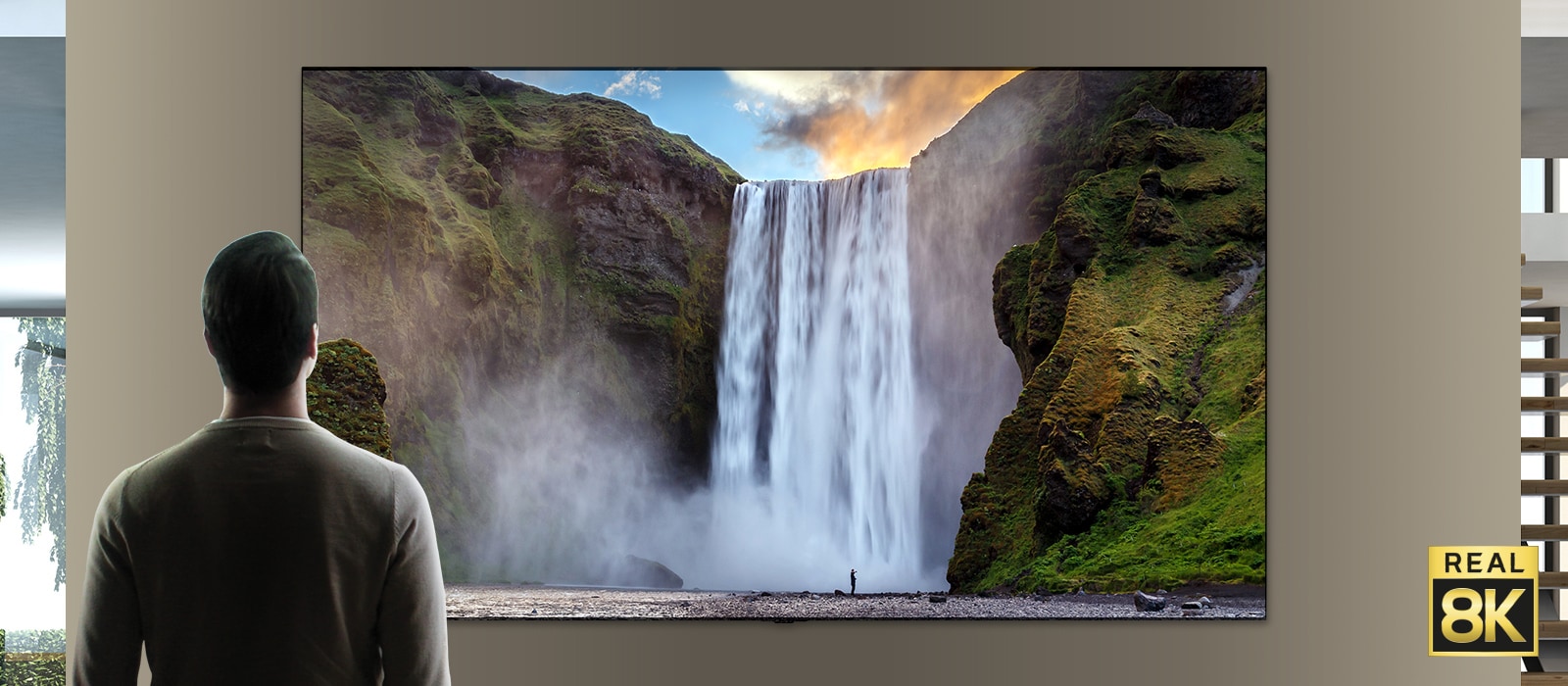 A man stood in front of an imposing view of a large waterfall crashing down cliffs. The scene zooms out to show the waterfall as an image on a wall mounted TV.