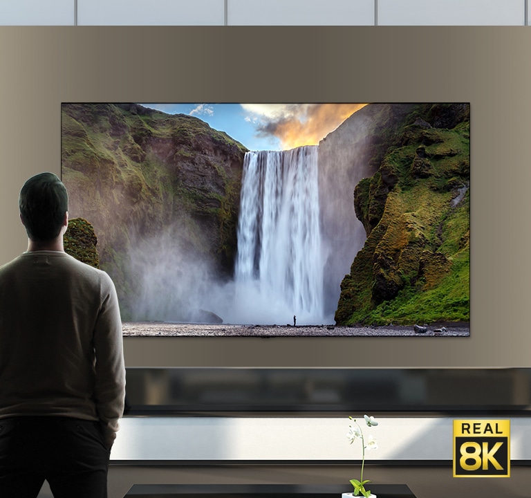 A man stood in front of an imposing view of a large waterfall crashing down cliffs. The scene zooms out to show the waterfall as an image on a wall mounted TV.