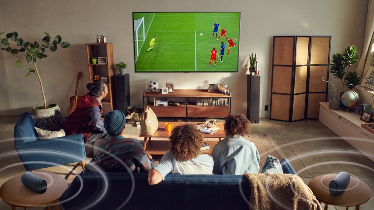 Five people sitting on the couch are watching a large screen TV displaying a soccer game with Bluetooth speakers making surrounding sound.